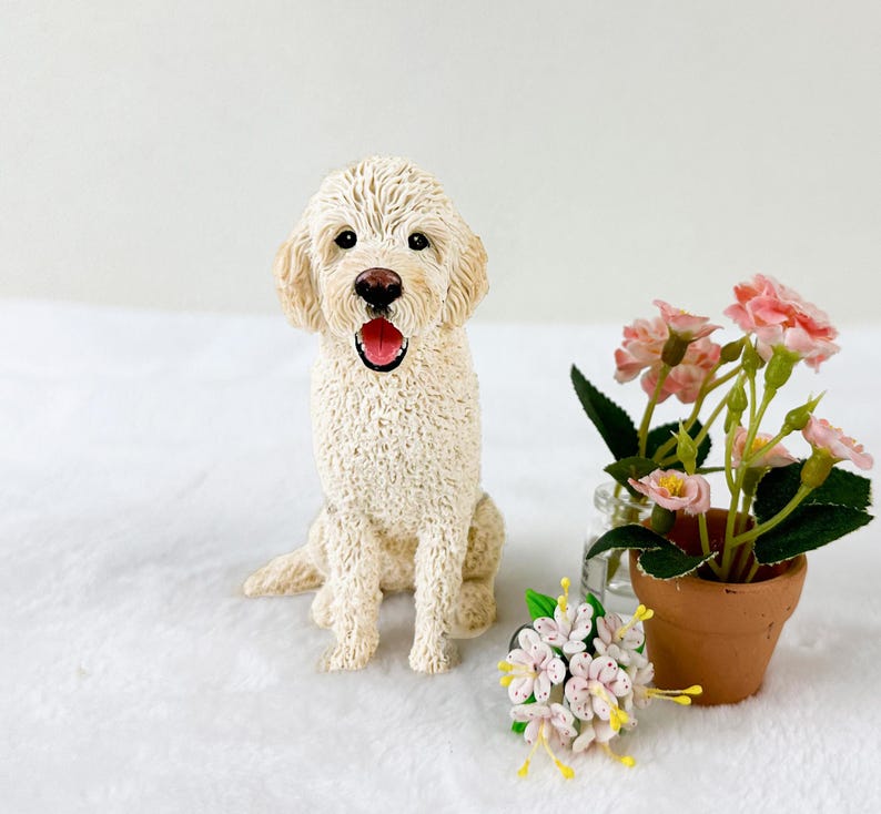 May include: A white cream-colored dog figurine with a brown nose and a pink tongue sticking out. The dog is sitting with its paws crossed and is looking directly at the camera. The figurine is on a white surface with a small bouquet of pink and white flowers in the foreground.