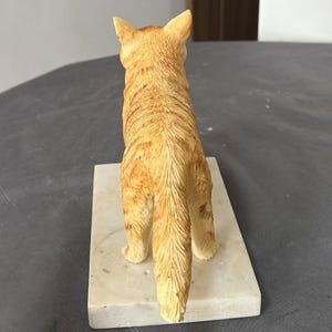 Puede incluir: Una figurita de un gato jengibre con una base de m&aacute;rmol blanco. El gato est&aacute; de pie sobre sus patas traseras con la cola enroscada detr&aacute;s de &eacute;l.
