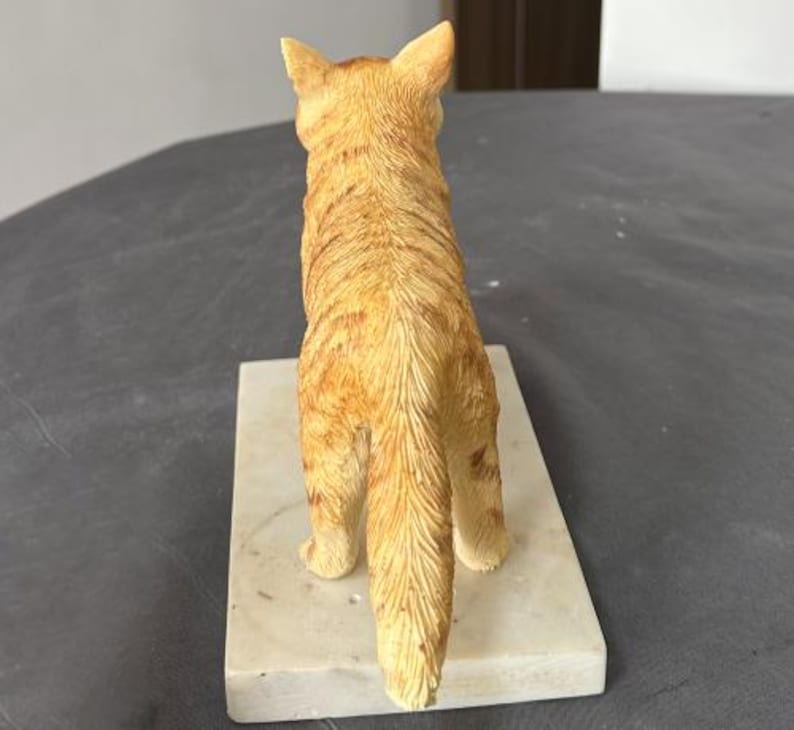 Puede incluir: Una figurita de un gato jengibre con una base de m&aacute;rmol blanco. El gato est&aacute; de pie sobre sus patas traseras con la cola enroscada detr&aacute;s de &eacute;l.