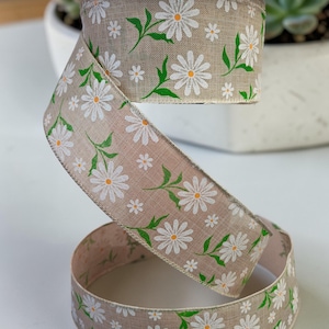 May include: A roll of beige ribbon decorated with a daisy pattern. The ribbon displays white daisies with yellow centres and green leaves. This ribbon is suitable for crafts, gift wrapping, or floral arrangements.