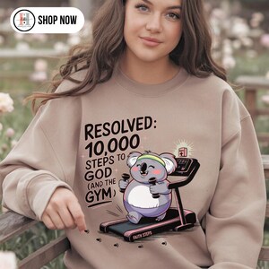 Puede incluir: Sudadera beige con el texto "RESOLVED: 10,000 STEPS TO GOD (AND THE GYM)". Un koala de dibujos animados está en una cinta de correr. La sudadera tiene cuello redondo y mangas largas.