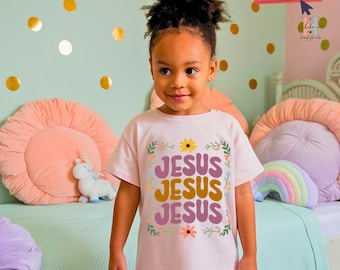 May include: A young child wearing a pink t-shirt with the words "Jesus Jesus Jesus" in a retro font and floral accents. The shirt is a light pink color and the text is in a yellow and brown color.