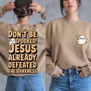 Puede incluir: Sudadera color beige con el texto "DON'T BE SPOOKED! JESUS ALREADY DEFEATED THE DARKNESS" en la espalda. Un pequeño gráfico de un fantasma sosteniendo una cruz está en la parte delantera. La sudadera es de estilo casual y cómodo.