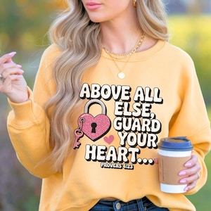May include: Mustard yellow sweatshirt with the text "ABOVE ALL ELSE, GUARD YOUR HEART..." and a pink heart-shaped lock and key graphic. The sweatshirt is paired with blue jeans and sunglasses.