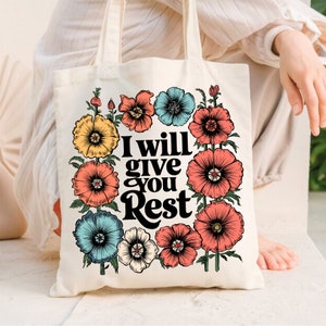 Pode incluir: Uma bolsa tote de lona branca com um design floral e o texto "I will give you Rest" em preto. As flores são em tons de rosa, amarelo, azul e branco.