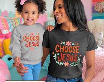 May include: A woman and a child wearing matching shirts with the text "CHOOSE JESUS" in orange and red, surrounded by floral designs. The woman's shirt is black, and the child's is gray. Both are smiling.