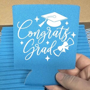 May include: A light blue can cooler with the words "Congrats Grad" in white script, along with a graduation cap and diploma graphic. The cooler is held by a hand and is part of a stack of similar coolers.