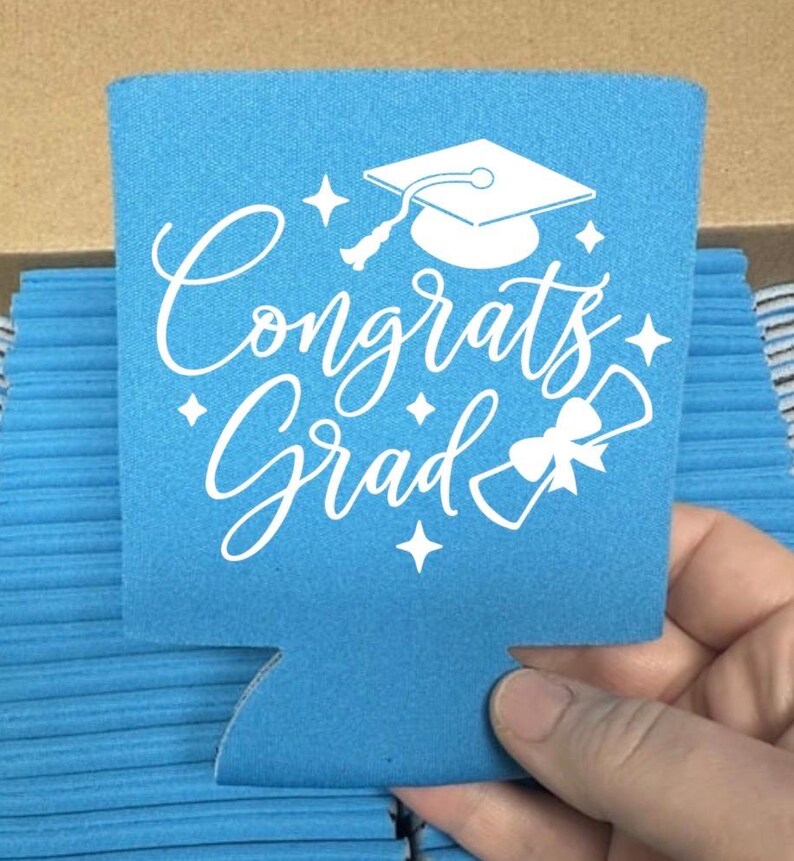 May include: A light blue can cooler with the words "Congrats Grad" in white script, along with a graduation cap and diploma graphic. The cooler is held by a hand and is part of a stack of similar coolers.