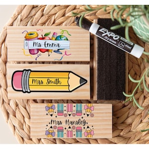 May include: Wooden teacher name stamps with colorful illustrations. One stamp features a pencil design with "Mrs. Smith" text. Another has school-themed graphics and "Ms. Emma" text. A third stamp has pencil and flower graphics with "Mrs. Hansley" text. An Expo dry erase marker and eraser are also present.