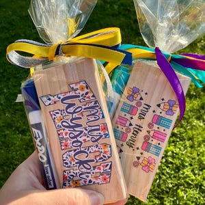 May include: Two gift bags with wooden teacher gifts. One has the text "Mrs Wysocki" in floral letters, the other says "Mrs. Henry" with pencil and bow graphics. Each bag is wrapped in clear cellophane and tied with colorful ribbons.