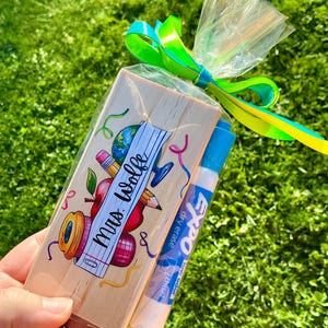 May include: A gift set for a teacher, including a wooden block with a school-themed design and the text "Mrs. Wolfe", and a blue and white Expo dry erase marker. The items are wrapped in clear cellophane and tied with a green and yellow ribbon.