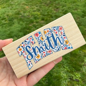 May include: A rectangular wooden block with the name "Smith" in a blue script font. The letters are outlined with a floral pattern in orange, red, and blue. The block is held against a background of green grass.
