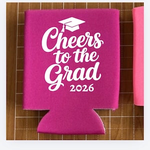 May include: A magenta-colored can cooler with the text "Cheers to the Grad 2026" in white script and a graduation cap graphic. The cooler is designed to hold a standard-sized can or bottle.
