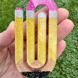 May include: A glittery pencil-shaped bookmark in yellow, silver, pink, and black. The bookmark has a pencil tip design with a yellow body and a pink top. The bookmark is held in a hand against a green background.