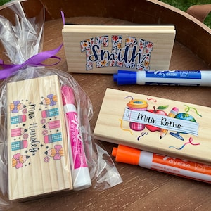 May include: Wooden dry erase board and eraser sets for teachers. One set is wrapped in cellophane with a purple ribbon. The boards are decorated with colorful school-themed graphics and teacher names. Includes dry erase markers.