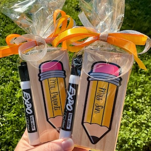 May include: Two gift bags with a pencil-shaped design, each containing a black Expo low odor chisel tip marker. The pencil design has a pink eraser and the words "Mr. Parenti" and "Mrs. Sin" on the pencils. The bags are tied with orange and silver ribbons.