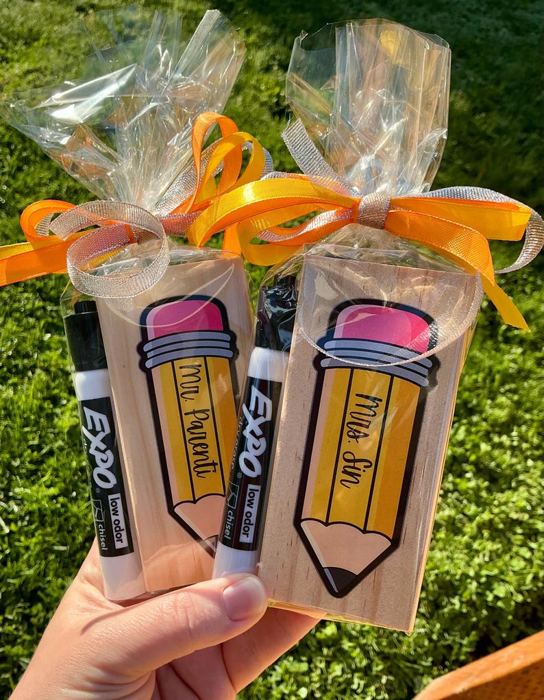 May include: Two gift bags with a pencil-shaped design, each containing a black Expo low odor chisel tip marker. The pencil design has a pink eraser and the words "Mr. Parenti" and "Mrs. Sin" on the pencils. The bags are tied with orange and silver ribbons.