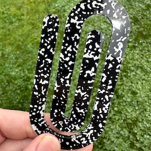 May include: A black and white patterned hair clip with a unique, abstract design. The clip is held against a backdrop of green grass. The pattern is reminiscent of a notebook paper design.