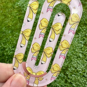 May include: A clear, U-shaped acrylic hair clip with a pink background. The clip is decorated with yellow bows and pencil illustrations. Small white flowers and gold stars are scattered across the pink background.
