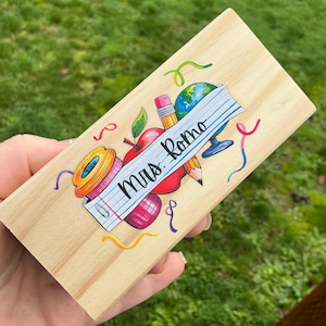 May include: A rectangular wooden plaque with a colorful teacher-themed design. The design includes a red apple, pencil, globe, and the text "Mrs. Romo" on a lined paper banner. The background is a natural wood grain.