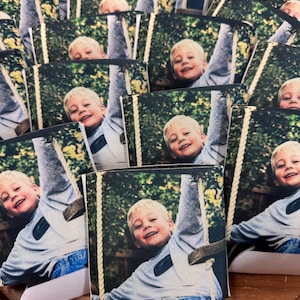 May include: A collection of beverage can coolers featuring a smiling child on a swing. The image shows the child with blond hair wearing a gray long-sleeve shirt and blue jeans. The background is a green, leafy outdoor setting.