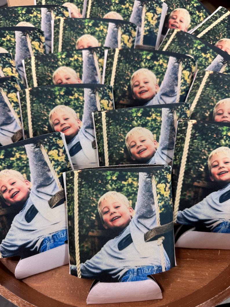 May include: A collection of beverage can coolers featuring a smiling child on a swing. The image shows the child with blond hair wearing a gray long-sleeve shirt and blue jeans. The background is a green, leafy outdoor setting.