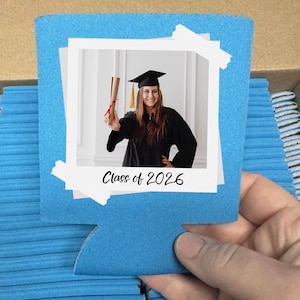 May include: A blue can cooler with a photo of a graduate in a cap and gown holding a diploma. The photo is framed with white borders and the text "Class of 2026" is below the photo. The cooler is held by a hand.