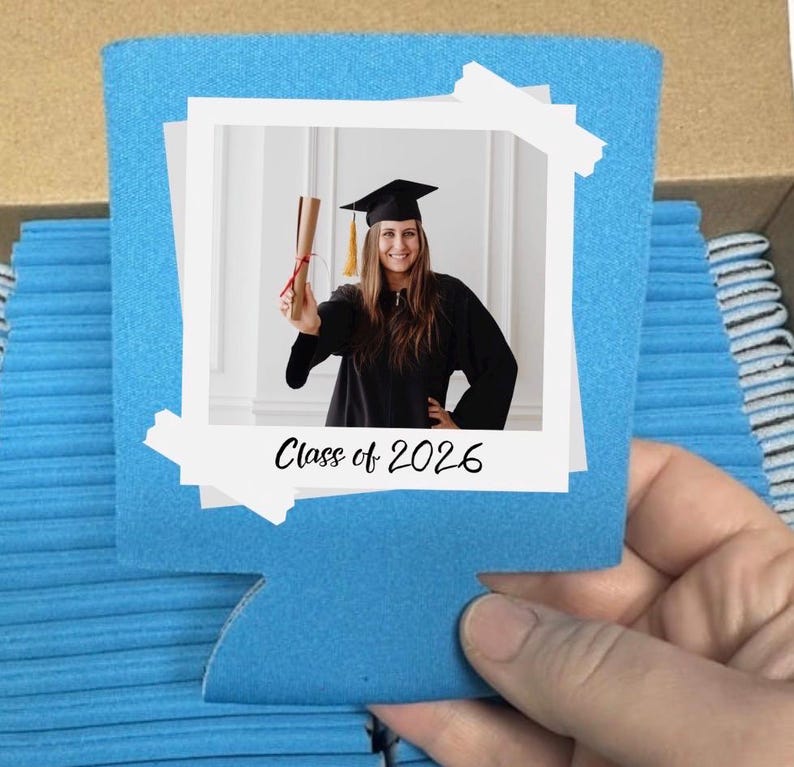 May include: A blue can cooler with a photo of a graduate in a cap and gown holding a diploma. The photo is framed with white borders and the text "Class of 2026" is below the photo. The cooler is held by a hand.