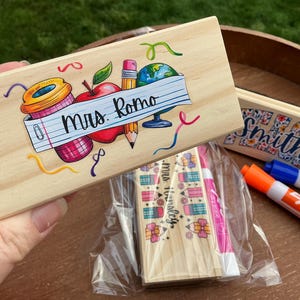 May include: Wooden box with a colorful school-themed design, including an apple, pencil, globe, and coffee cup. The box is labeled "Mrs. Romo" in black script. Other boxes and dry erase markers are also visible.