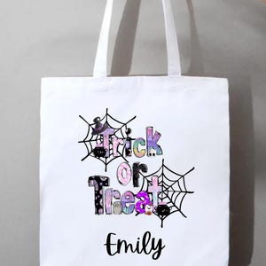 May include: White tote bag with the words "Trick or Treat" in colorful, patterned letters, framed by black spiderwebs. The name "Emily" is printed below the text in a cursive font. The bag has white handles.