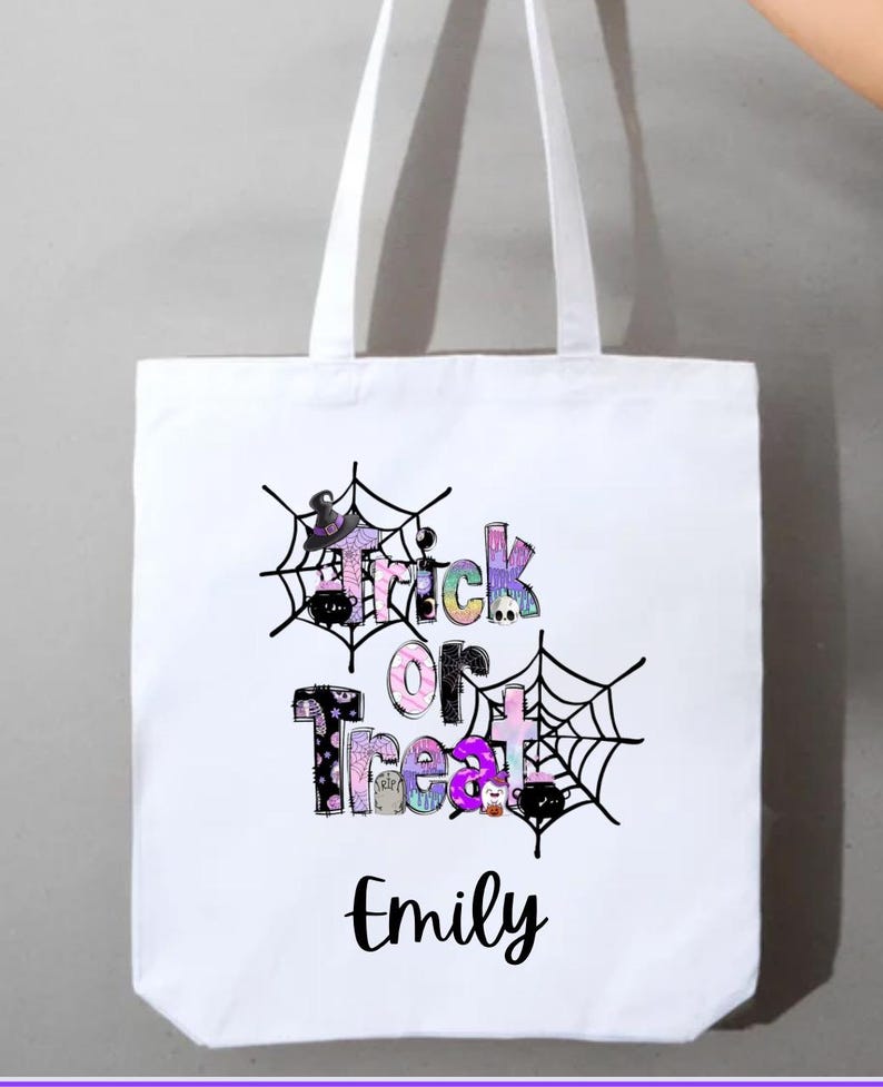 May include: White tote bag with the words "Trick or Treat" in colorful, patterned letters, framed by black spiderwebs. The name "Emily" is printed below the text in a cursive font. The bag has white handles.