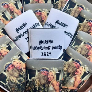 May include: A collection of white can coolers with the text "Marine Halloween party 2024" in black script. Each cooler has a photo of a smiling couple. The coolers are arranged in a wooden container.