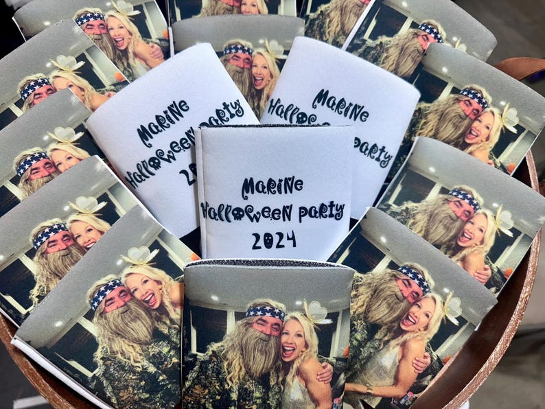 May include: A collection of white can coolers with the text "Marine Halloween party 2024" in black script. Each cooler has a photo of a smiling couple. The coolers are arranged in a wooden container.