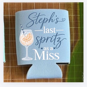 May include: A light blue can cooler with the text "Steph's - last spritz as a Miss" in script and block letters. A graphic of a cocktail with an orange slice and a straw is also featured.