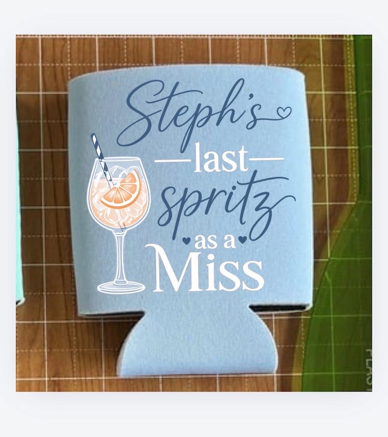 May include: A light blue can cooler with the text "Steph's - last spritz as a Miss" in script and block letters. A graphic of a cocktail with an orange slice and a straw is also featured.