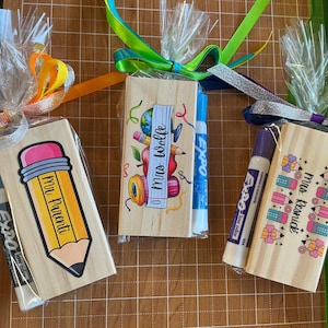 May include: Three teacher gifts wrapped in cellophane with colorful ribbons. Each gift includes a wooden block with a teacher-themed design and an Expo marker. One block says "Mr. Parenti" with a pencil design.