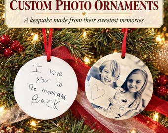 May include: Two round white ceramic ornaments hanging by red ribbons. One ornament features black handwritten text that reads I love You To The moon and Back. The other ornament displays a black and white photograph of two children holding a drawing.