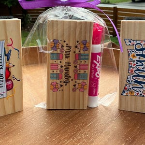 May include: Wooden dry erase board erasers with colorful designs and text. One eraser has a school-themed design with the text "Mrs. Romo", another has the text "Mrs. Hansley", and the third has the name "Smith" in a floral pattern. Includes dry erase markers.