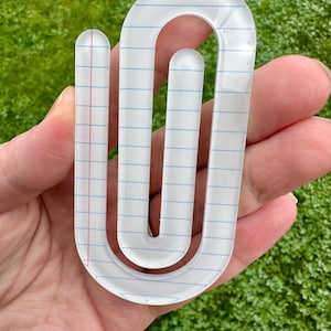May include: A clear, oversized paperclip-shaped object with a notebook paper design. The paperclip has blue horizontal lines and a red vertical margin line. The object is held in a hand against a green, blurred background.