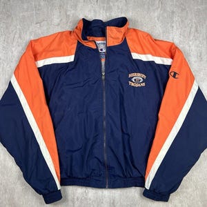 May include: A blue, white, and orange Champion brand jacket with a zipper closure. The jacket has a white stripe and orange stripe on each sleeve. The jacket has the text "Hershey Trojans" embroidered on the front.