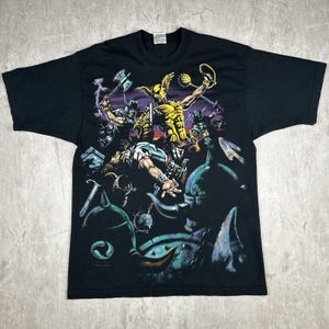 Op de afbeelding: Zwart T-shirt met een grafische print van een gevleugelde krijger in geel en groen die vecht tegen andere krijgers. De grafiek is gedetailleerd en bevat bliksemschichten en andere symbolen.