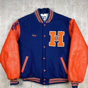 May include: A blue and orange varsity jacket with leather sleeves. The jacket has a large "H" on the front with a mascot inside the letter. The name "Wade" is embroidered on the left side of the jacket.