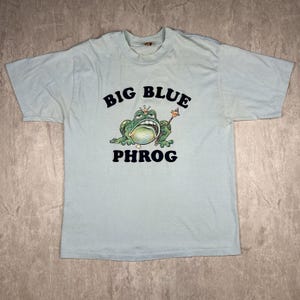 May include: A light blue t-shirt with a cartoon frog wearing a crown and holding a scepter. The text "BIG BLUE PHROG" is printed on the shirt.