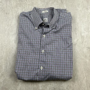 以下が含まれることがあります： 白、黒、紫のチェック柄のボタンダウン長袖シャツ。シャツの左側に小さなポケットがあり、前面には「The English Shop」というブランドラベルが付いています。