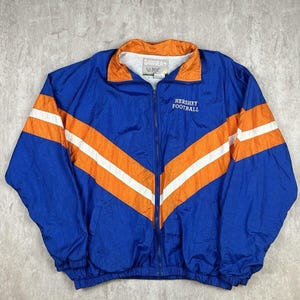 May include: A blue and orange windbreaker jacket with white stripes. The jacket has a zipper closure and a  "Sahara" and "Hershey Football" logo.