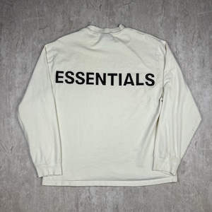 Op de afbeelding: Crèmekleurig shirt met lange mouwen met het woord "ESSENTIALS" in grote, zwarte blokletters op de rug. Het shirt heeft een ronde hals en lange mouwen. De stof lijkt een zacht, comfortabel materiaal te zijn.