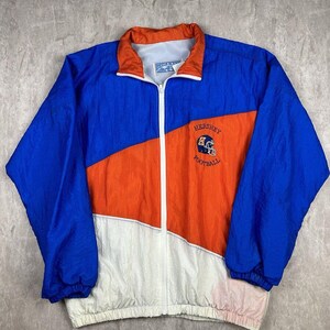 May include: A blue, orange, and white windbreaker jacket with a zipper closure. The jacket has a logo on the front that says "Hershey Football" with a football helmet graphic.