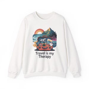 Könnte beinhalten: Weißes Sweatshirt mit einem farbenfrohen Reise-Motiv. Das Design zeigt einen Koffer, einen Strand, Berge und einen Regenbogen, mit dem Text "Travel is my Therapy".
