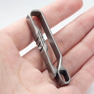 Könnte beinhalten: Ein silberfarbener Metallclip mit U-förmigem Design. Der Clip hat eine Reihe diagonaler Rillen auf einer Seite und eine kleine sechseckige Öffnung unten. Der Clip wird in der Hand einer Person gehalten.