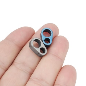 Op de afbeelding: Twee titanium paracord clips, één in een blauwgrijze kleur en één in een zilveren kleur. De clips hebben twee gaten om paracord aan te bevestigen.